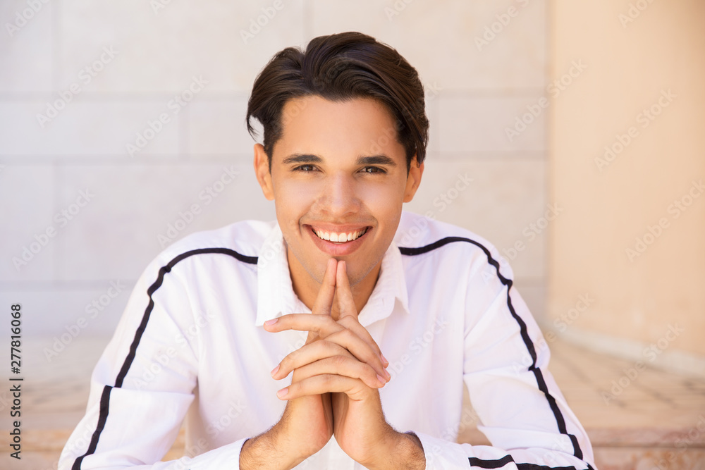 Happy Latin man keeping hands together and sitting outdoors. Handsome young guy wearing shirt and looking at camera. Man portrait concept. Front view. Happy Latin man keeping hands together and sitting outdoors. Handsome young guy wearing shirt and looking at camera. Man portrait concept. Front view.
