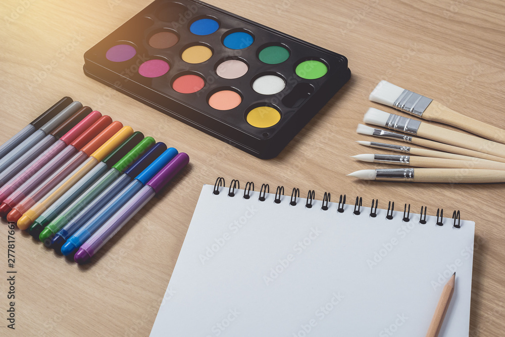 Obraz premium Notepad or notebook with Many colorful pens, paintbrush and Watercolor palette on brown wood table.using for Arts and Education background