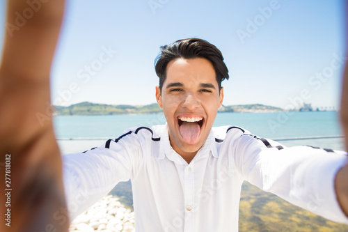 Happy Latin man grimacing and taking selfie photo at seaside. Young man showing tongue and holding unseen camera with sea and blue sky in background. Tourism and selfie concept. Front view.