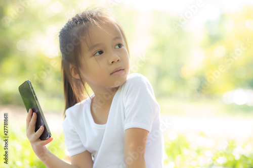 Beautiful asian child smiling using smart mobile phone in the garden, kid have passion playing game digital on smartphone on internet network in the park with happy, lifestyle and technology concept.