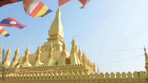 (Selective focus) Slow motion video of Laos flags waving in the foreground with the beautiful Pha That Luang in the background. Pha That Luang is a gold-covered large Buddhist stupa in Vientiane, Laos