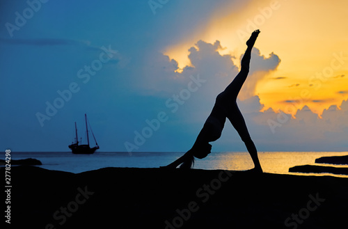 Silhouette of a young woman doing yoga exercises by the sea against the backdrop of colourful dawn 