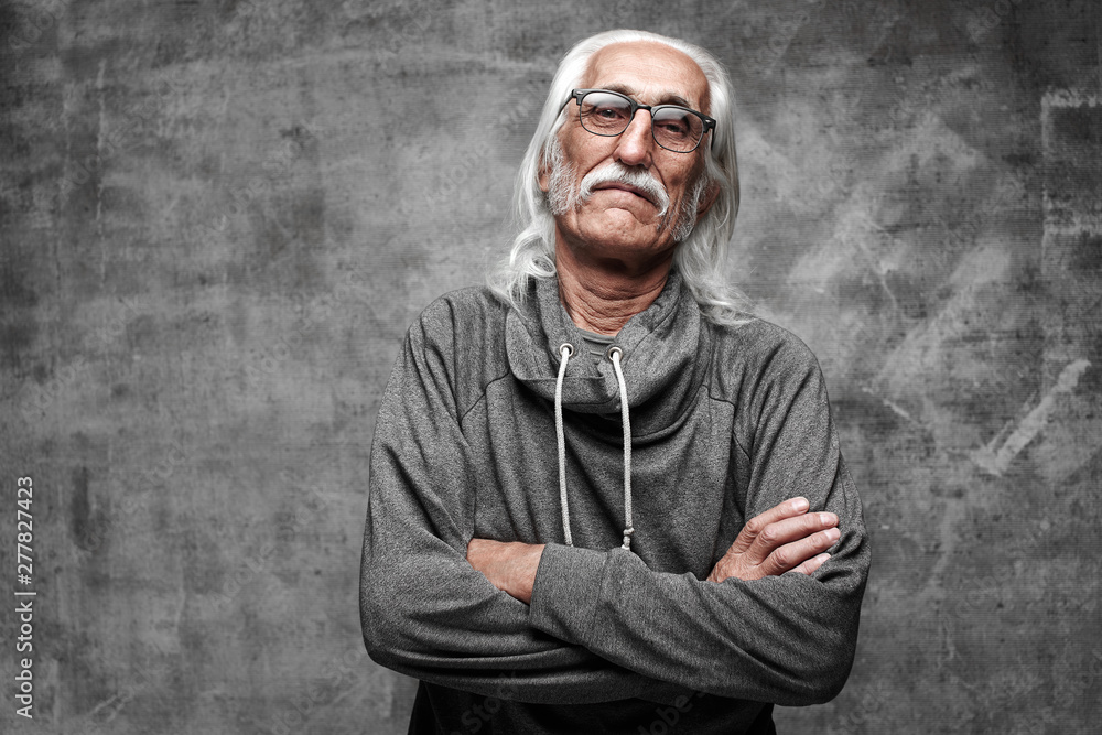 Old Caucasian gray-haired cool grandfather with glasses incredulously looks into the camera with crossed arms and serious face with copy space in studio Old Caucasian gray-haired cool grandfather with glasses incredulously looks into the camera with crossed arms and serious face with copy space in studio