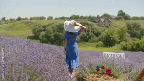 Beautiful healthy woman in trendy dress and sun hat running joyfully through lavender field while enjoying outdoor leisure in countryside. Carefree younng female relaxing on summer vacations in nature photo