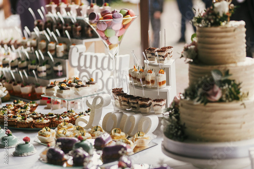 Delicious candy bar at wedding reception. White and chocolate desserts with fruits, macarons, cake, cupcakes on stand, modern sweet table at wedding or baby shower. Luxury catering concept