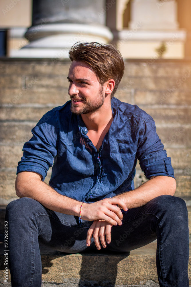 Fototapeta premium One handsome young man in urban setting in summer day, wearing blue shirt, sitting on stone steps
