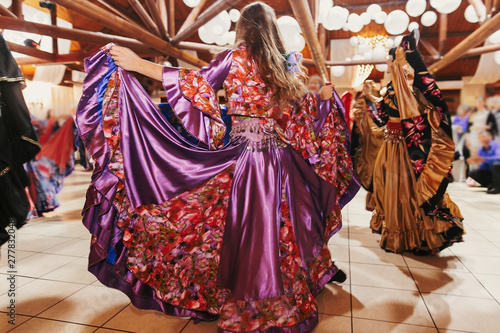 Beautiful gypsy girls dancing in traditional purple floral dress at wedding reception in restaurant. Woman performing romany dance and folk songs in national clothing. Roma gypsy festival