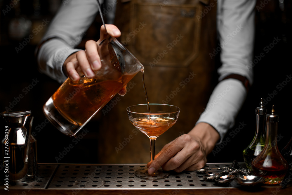 Professional male bartender pouring a brown alcoholic cocktail from the measuring cup to the glass