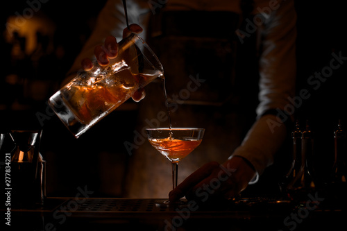 Male bartender pouring a brown alcoholic cocktail from the measuring cup to the glass in the dark