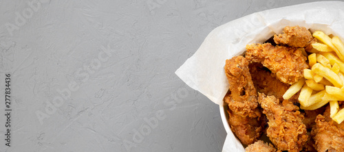 Fastfood: fried chicken drumsticks, spicy wings, French fries and chicken strips in a paper box over gray background, top view. Space for text. photo