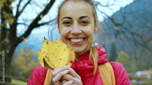 young pretty smiling woman in red jacket standing outdoors, holding a few dry yellow leaves in hands and plays with them, playfully hiding behind leaves and peeking out photo