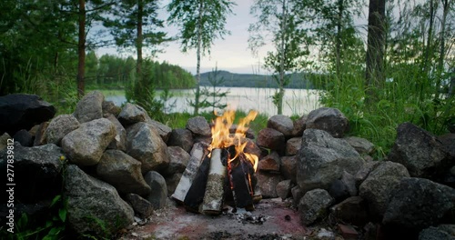 Cinematic shot of moody campfire on beautiful evening, camera sliding forward, lake view background