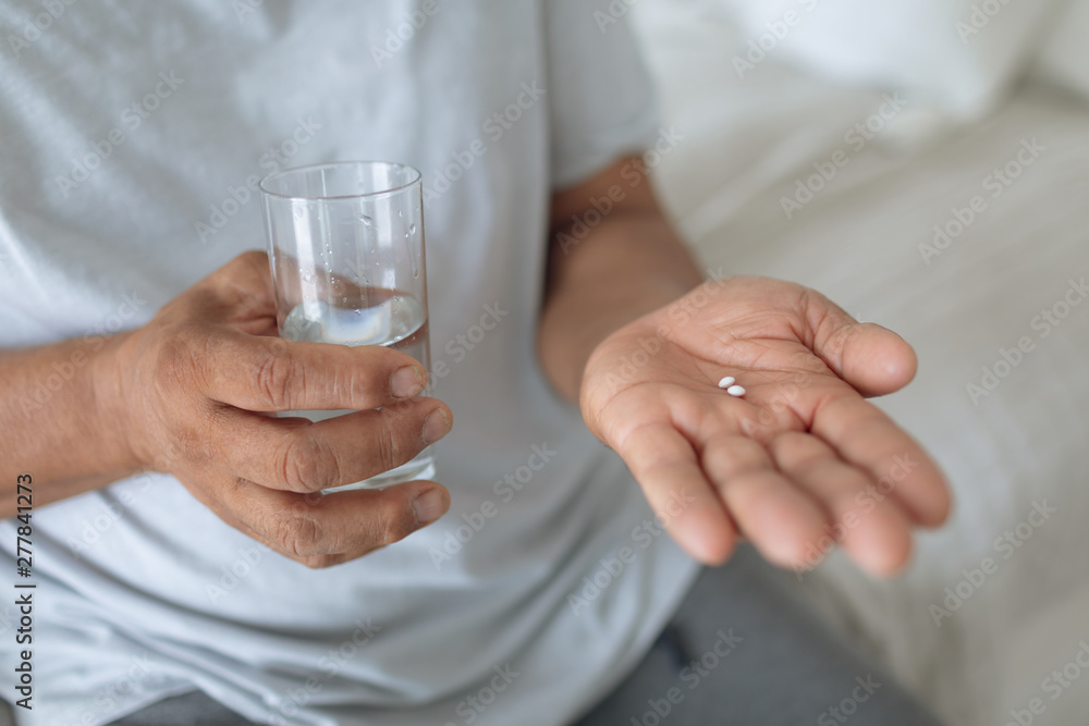 Man sitting on the bed while holding pills and a glass of water Man sitting on the bed while holding pills and a glass of water