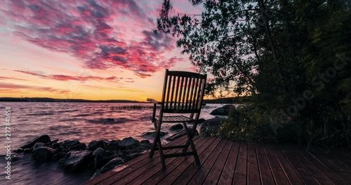 Beautiful sliding time-lapse of a colorful sunset over the lake, chair on foreground, wind is blowing and waves hit the coastline.