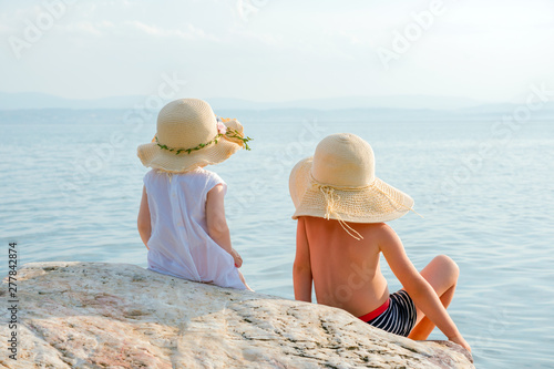 Back view of two kids sitting on stone and looking to ocean. Little travelers near the ocean. Boy and girl in summer hats, panamas, on seashore. Summer holidays concept. Traveling background photo