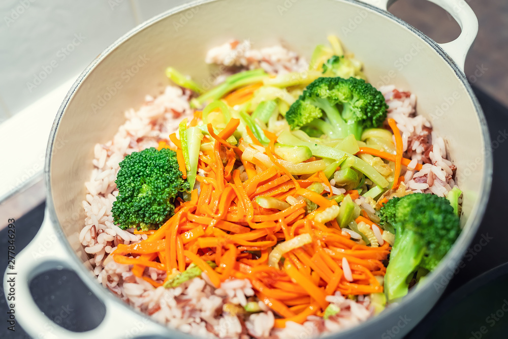 Brown rice with organic unmixed peeled sliced vegetables cooking in cast iron saucepan. Bright broccoli, carrot and zucchini over cereal background. Vegetarian healthy food Brown rice with organic unmixed peeled sliced vegetables cooking in cast iron saucepan. Bright broccoli, carrot and zucchini over cereal background. Vegetarian healthy food