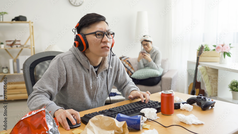 Lazy asian guy playing online video game on computer while his girlfriend  cleaning table at home. frowning woman murmur at her boyfriend. man in  headphones concentrated having fun relax on technology Stock