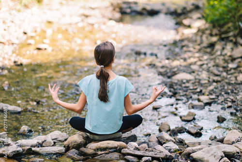 Young girl yoga meditating at sunrise on the shore of a mountain stream. Teenage model meditating in peaceful harmony