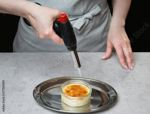 Obraz na plátně An image of a creme brulee in a transparent glass bowl on an iron tray; a woman