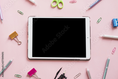 Digital tablet surrounded by various stationeries on pink background