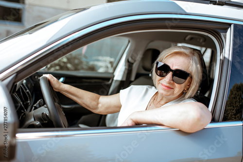 Portrait of a senior woman driver sitting in the modern car, looking out the window. Concept of an active people during retirement age