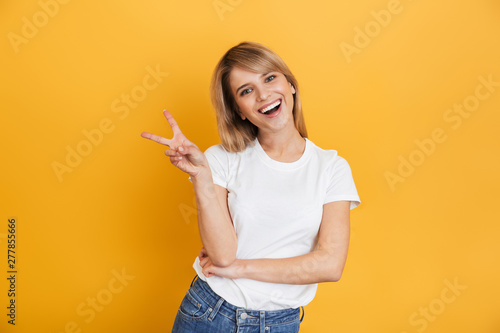 Happy young pretty blonde woman posing isolated over yellow wall background dressed in white casual t-shirt showing peace gesture.