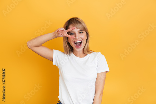 Optimistic emotional young pretty blonde woman posing isolated over yellow wall background dressed in white casual t-shirt showing peace gesture.