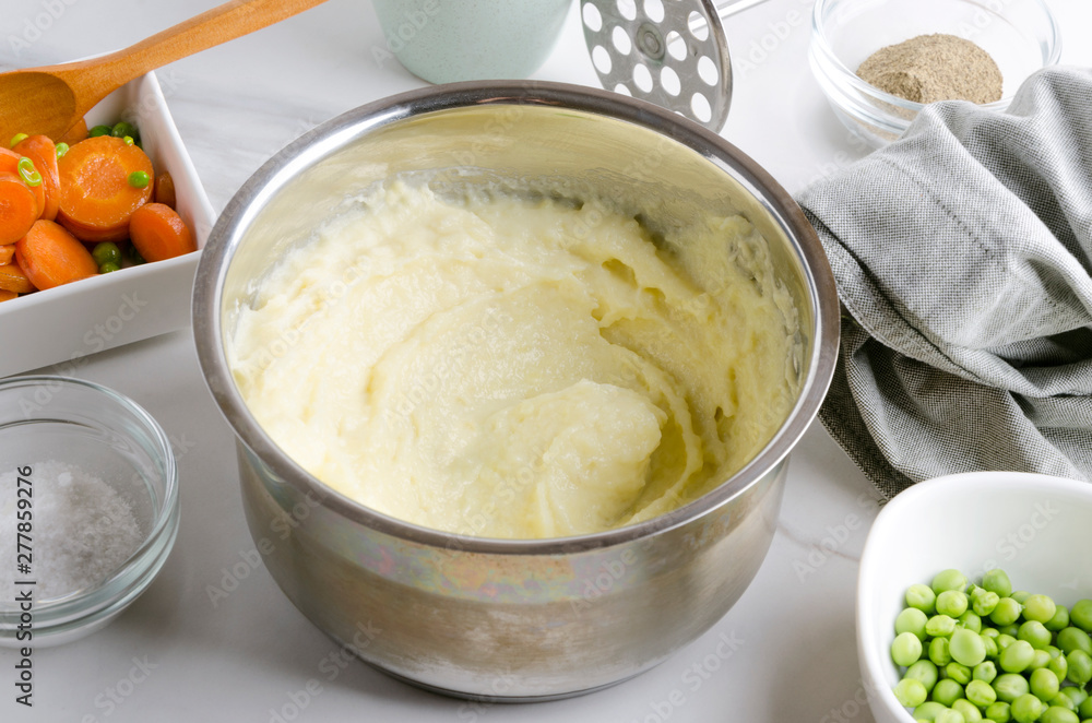 Delicious fresh cooked mashed potatoes in the pot, vegetables and spices on the white table Delicious fresh cooked mashed potatoes in the pot, vegetables and spices on the white table