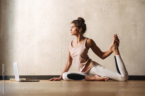 Photo of young concentrated woman doing yoga exercises using laptop while sitting on floor at home