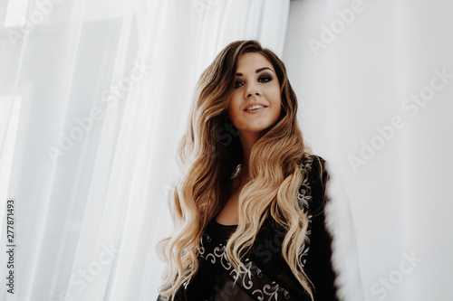 Beautiful sexy brown curly long hair woman posing in the camera. Studio shooting in interior. Model in bed. Happy glamorous woman