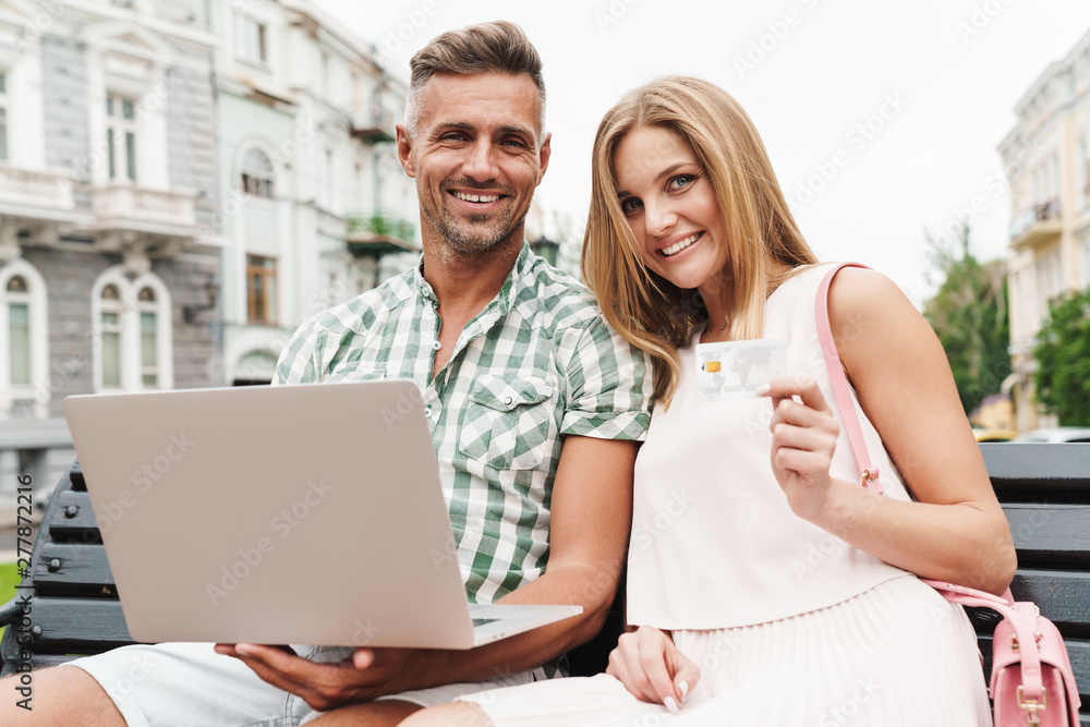 Portrait of caucasian young couple holding credit card and using laptop while sitting on bench in city street