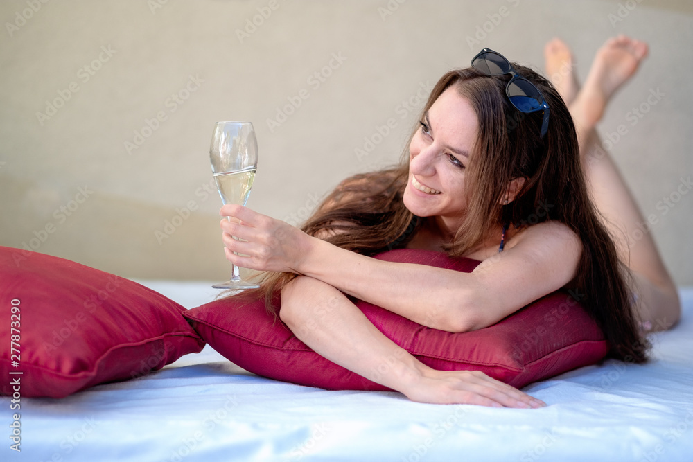 Laughing young sexy woman in bikini lies with a glass of sparkling wine in the lounge and gives a toast Laughing young sexy woman in bikini lies with a glass of sparkling wine in the lounge and gives a toast