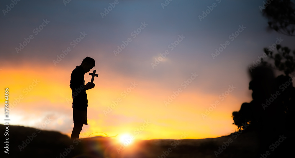 Silhouette of young male christian standing and holding a cross for blessing from god with light of sunset background, christian hope concept.