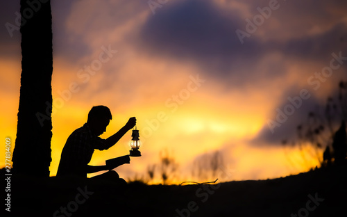 Silhouette of young male christian sitting holding oil lamp and reading bible with light of sunset background  christian hope concept.