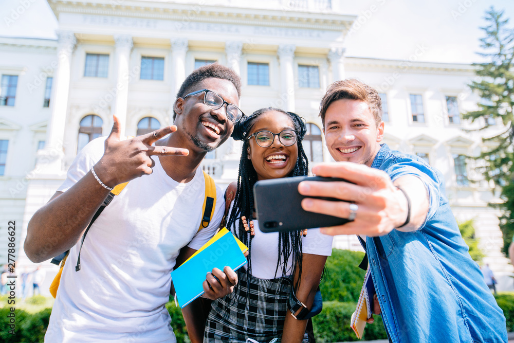 Obraz premium Happy interracial boys and african girl friend posing for cute selfie.Young friends in t-shirts photographing together against university building. Relationship multhetnic friendship concept.