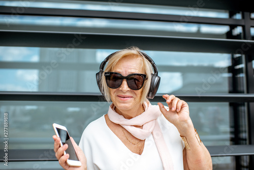Lifwstyle portrait of a stylish senior woman listening to the music with headphones and phone standing on the modern building background