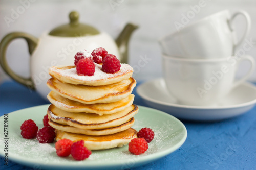 stack of baked homemade pancakes with raspberries, sprinkled with powdered sugar and tea utensils in the background, selective focus