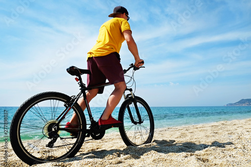 Young man with fit body riding the mtb mountain bike on sandy beach with beautiful azure water sea view. Muscle male wearing bright yellow t-shirt cycling on ocean shore. Close up, copy space.