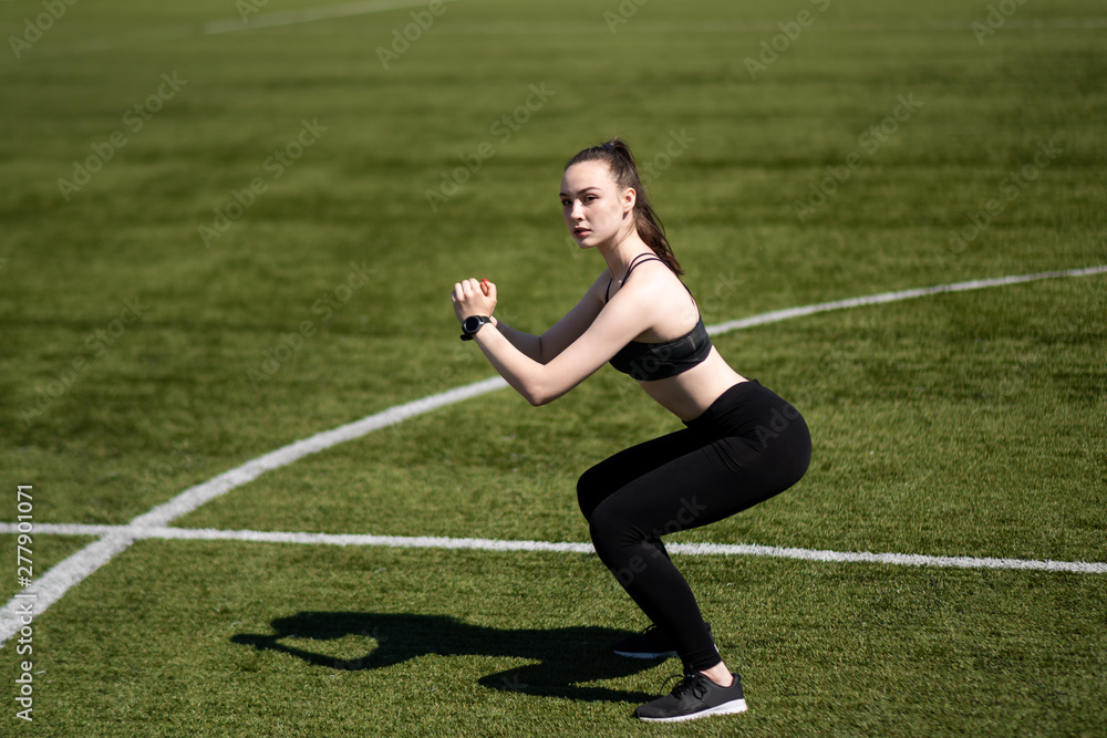 Girl doing a squat at the stadium. Black leggings and top. Football field.