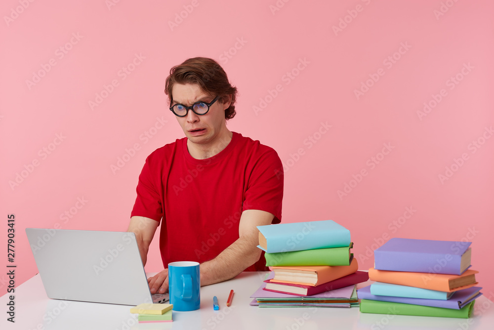 Portrait of shocked young student in glasses wears in red t-shirt, man sits by the table and working with laptop and books, looks at the monitor with disgusted, isolated over pink background. Portrait of shocked young student in glasses wears in red t-shirt, man sits by the table and working with laptop and books, looks at the monitor with disgusted, isolated over pink background.