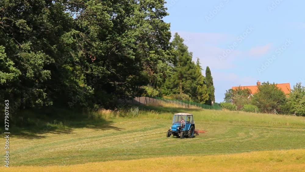 Blue tractor turn dry grass, hay near forest, Rural scene, Czech landscape Blue tractor turn dry grass, hay near forest, Rural scene, Czech landscape