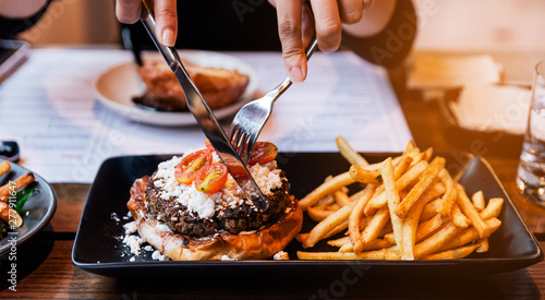 Hand fork pricking Cheeseburger with grilled beef, feta cheese and sliced tomato served with fries in black plate.