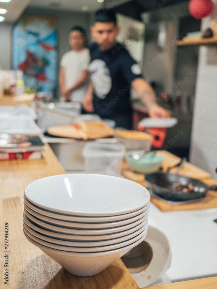 Stack of white bowls prepared for serving ramen on wooden counter and blurred male cooks in kitchen Stack of white bowls prepared for serving ramen on wooden counter and blurred male cooks in kitchen