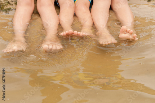 Funny kids in bathing suits sit together on sand with their legs in water. boys are happy on holiday in village together. Summer day, river, swimming in the water. Close-up legs and feet