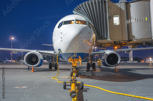 Passenger airplane is parked connected by a ladder at the terminal building in the airport, near the front trailer winch for tow tractor at night.