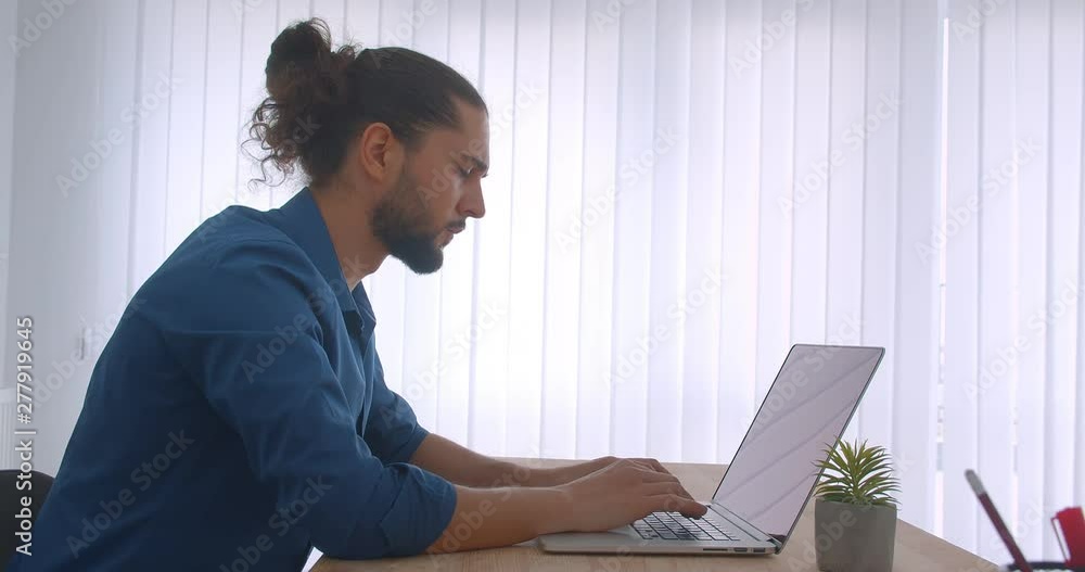 Profile shot of progressive businessman with ponytail working with laptop in light office. Profile shot of progressive businessman with ponytail working with laptop in light office.