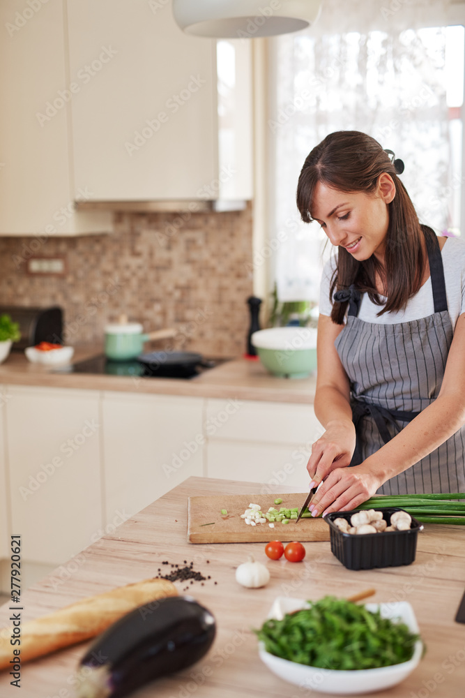 Smiling Caucasian female chef in apron chopping leek in kitchen and preparing dinner. Preparation of domestic food concept. Smiling Caucasian female chef in apron chopping leek in kitchen and preparing dinner. Preparation of domestic food concept.