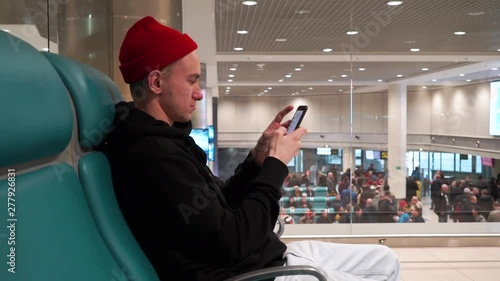 The young man is tapping number on mobile phone standing next to the information board at the airport. Attractive man in red hat talking on mobile phone.