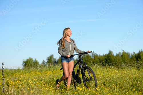 The caucasian young sportwoman with her bicycle is going on the blossoming meadow in summer day.