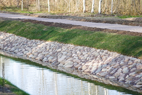 Natural stone paved shores of the canal in the park. Green grass on the banks. The sky is reflected in the channel. Place for jogging walks and outdoor recreation.